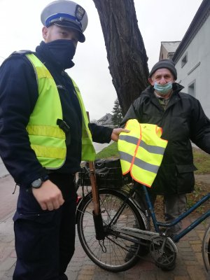 Zdjęcie przedstawia umundurowanego policjanta ruchu drogowego który przekazuje starszemu mężczyźnie, rowerzyście kamizelkę odblaskową.